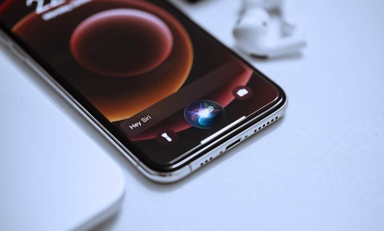 iPhone screen with Siri, showing "Hey Siri." AirPods are nearby.