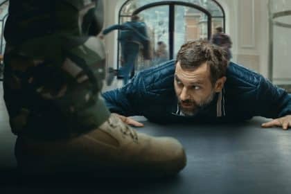 A man lies on the ground inside a building while people run in the background, suggesting a tense standoff scene.