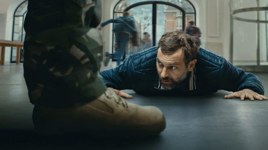 A man lies on the ground inside a building while people run in the background, suggesting a tense standoff scene.