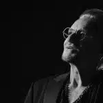 Bono in a black-and-white portrait wearing glasses and a dark jacket.