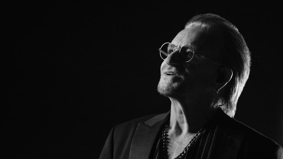 Bono in a black-and-white portrait wearing glasses and a dark jacket.