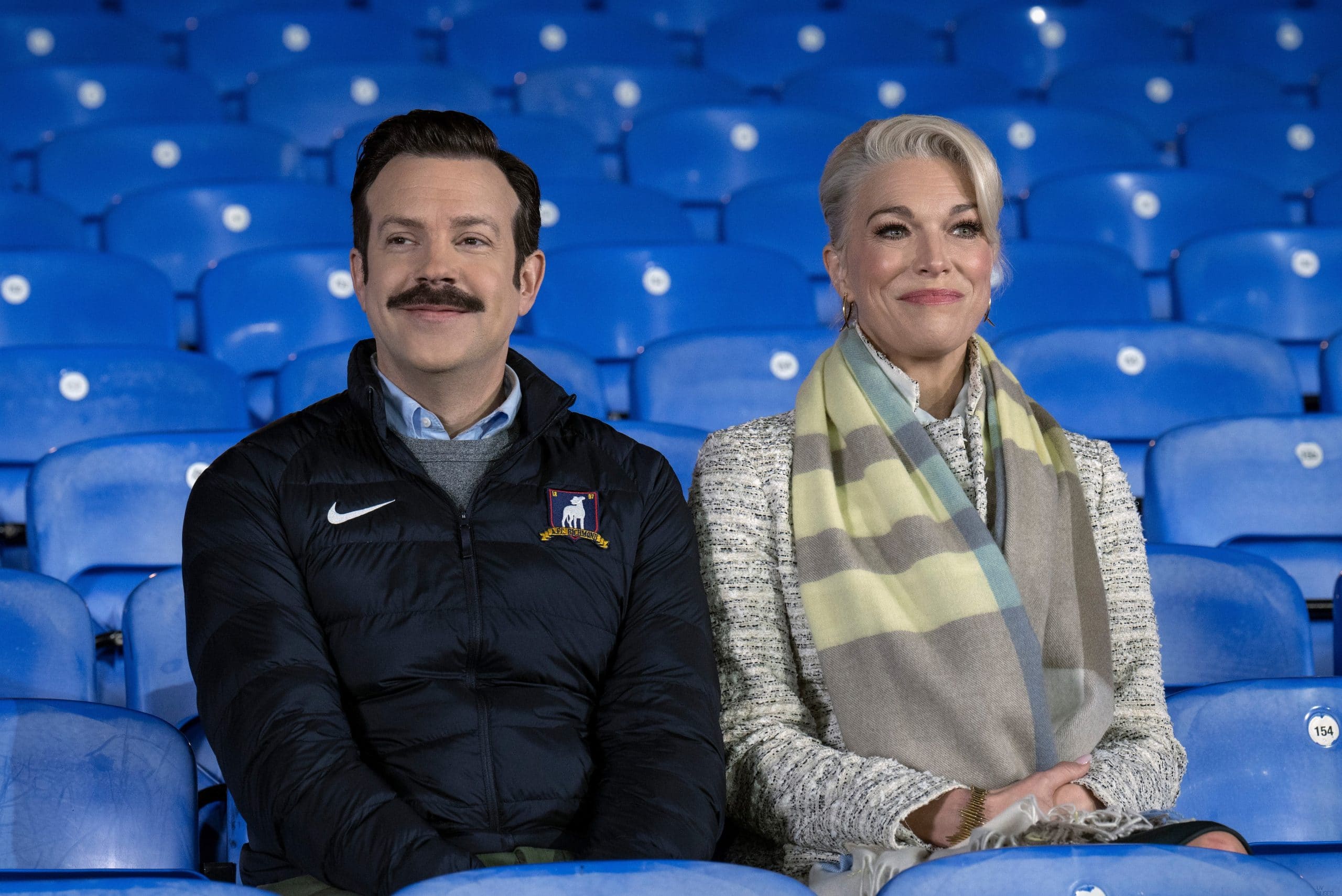 Jason Sudeikis and Hannah Waddingham sitting in stadium seats during "Ted Lasso" scene.