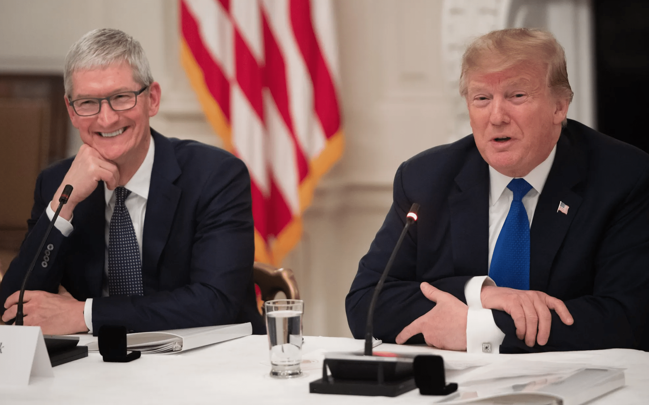 Tim Cook and Donald Trump seated at a meeting, speaking with microphones.