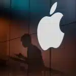 A glowing Apple logo on a dark building wall with a shadow of a person using a phone.
