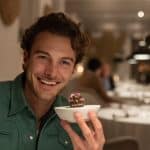 Man smiling and holding a small gourmet dish in a fine dining restaurant.