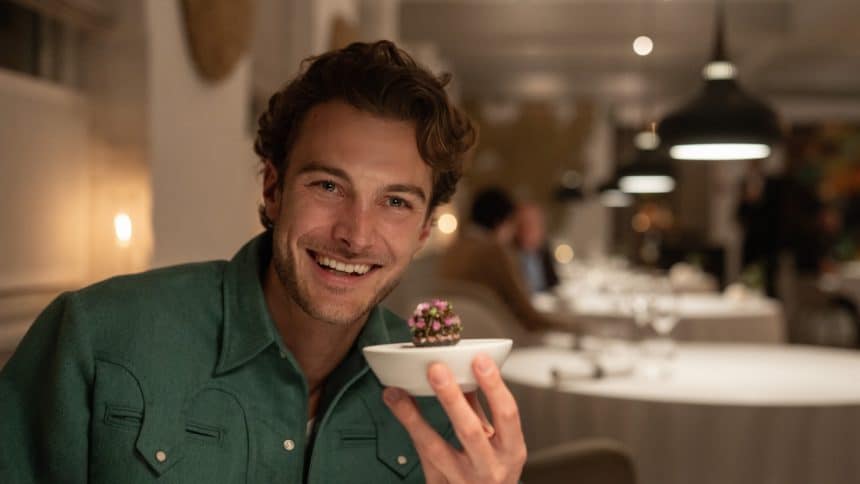 Man smiling and holding a small gourmet dish in a fine dining restaurant.
