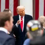 Donald Trump speaking to a crowd in front of a U.S. flag backdrop.