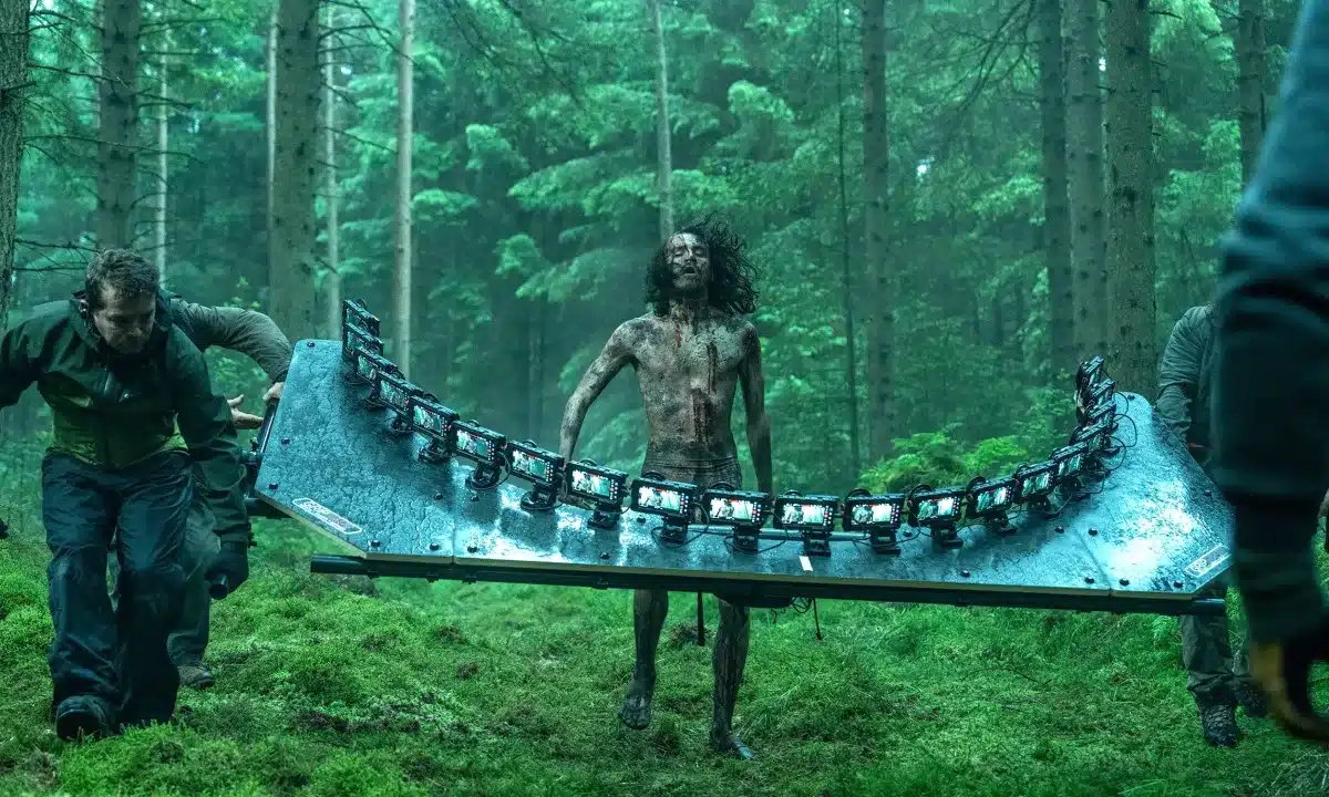 Film crew carries a curved rig of iPhones past a bloodied actor in a forest.