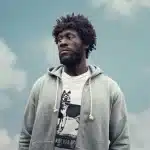 Stormzy standing outdoors against a cloudy sky, wearing a gray hoodie and a graphic T-shirt.