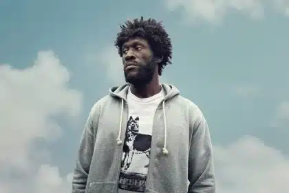 Stormzy standing outdoors against a cloudy sky, wearing a gray hoodie and a graphic T-shirt.