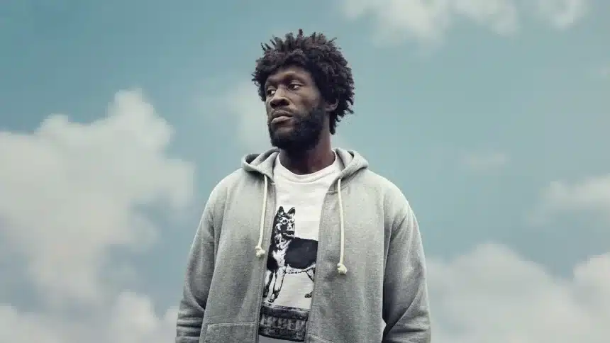 Stormzy standing outdoors against a cloudy sky, wearing a gray hoodie and a graphic T-shirt.