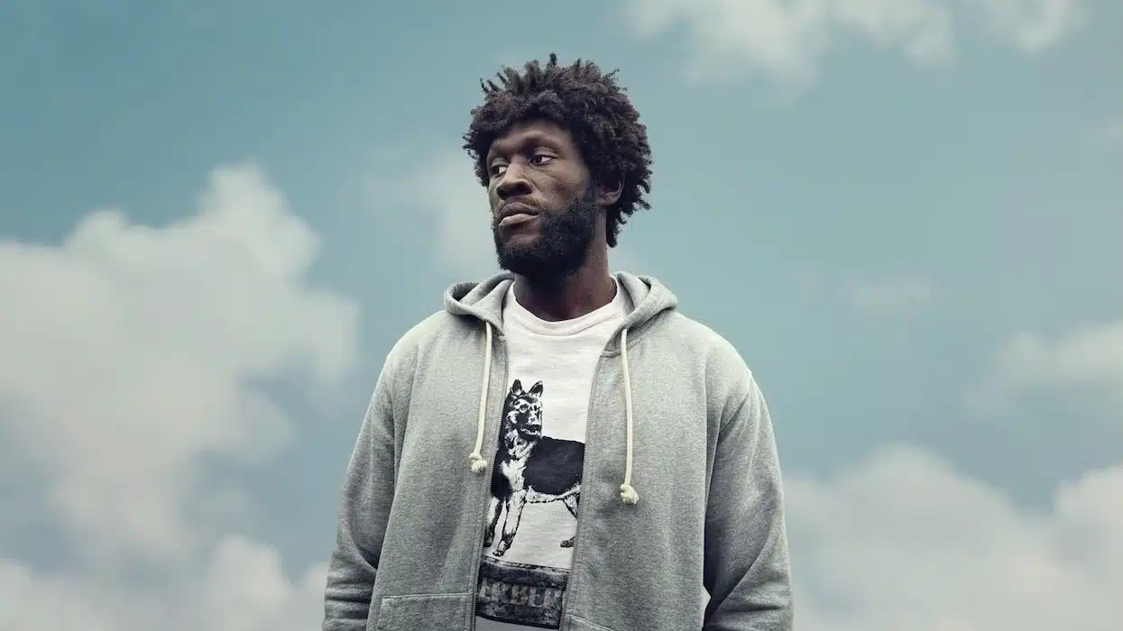 Stormzy standing outdoors against a cloudy sky, wearing a gray hoodie and a graphic T-shirt.