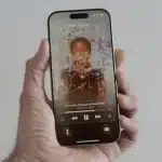 Hand holding an iPhone showing animated album art and music controls on the lock screen.