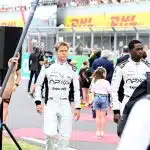 Brad Pitt and co-star in racing suits walking on an F1 track set with crew members around.