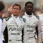Brad Pitt and co-star in racing suits walking on an F1 track set among crew members.