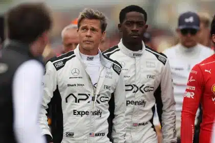 Brad Pitt and co-star in racing suits walking on an F1 track set among crew members.