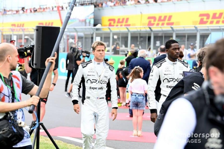 Brad Pitt and co-star in racing suits walking on an F1 track set with crew members around.