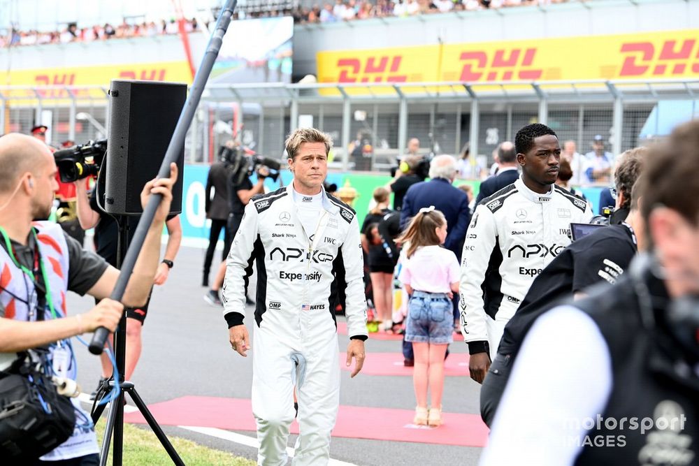 Brad Pitt and co-star in racing suits walking on an F1 track set with crew members around.
