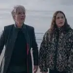 Two women stand on a beach looking concerned, with waves and cliffs in the background.