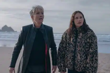 Two women stand on a beach looking concerned, with waves and cliffs in the background.