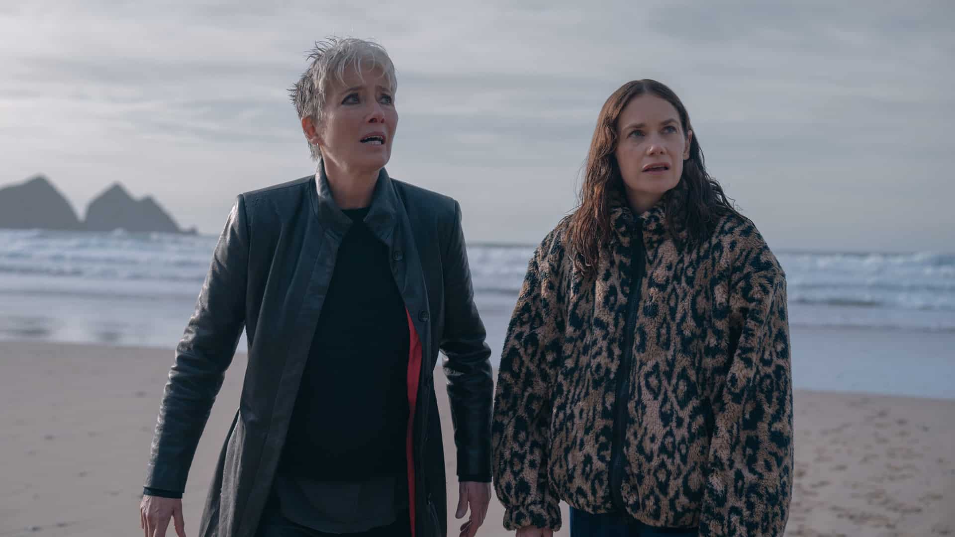 Two women stand on a beach looking concerned, with waves and cliffs in the background.