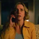 A woman in a yellow jacket looks shocked while talking on the phone.