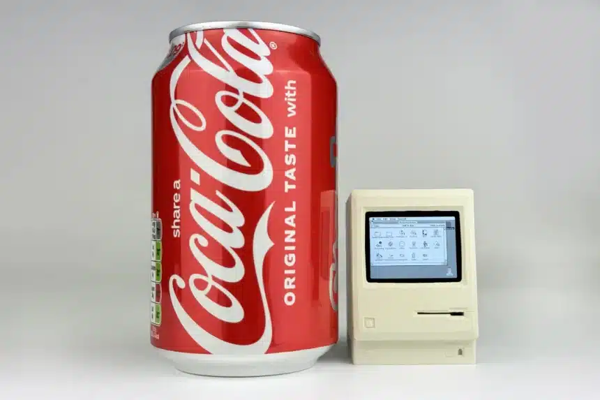 A tiny Macintosh replica next to a Coca-Cola can for size comparison.