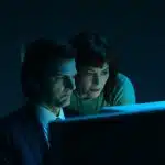 Two characters from Severance look at a glowing computer screen in a dark room.