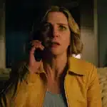 A woman in a yellow jacket looks tense while speaking on the phone in a dimly lit room.
