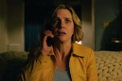 A woman in a yellow jacket looks tense while speaking on the phone in a dimly lit room.