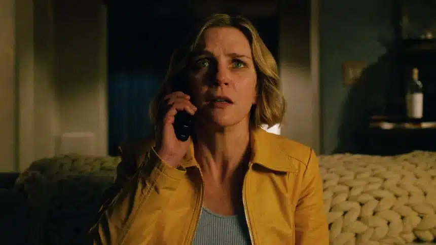 A woman in a yellow jacket looks tense while speaking on the phone in a dimly lit room.