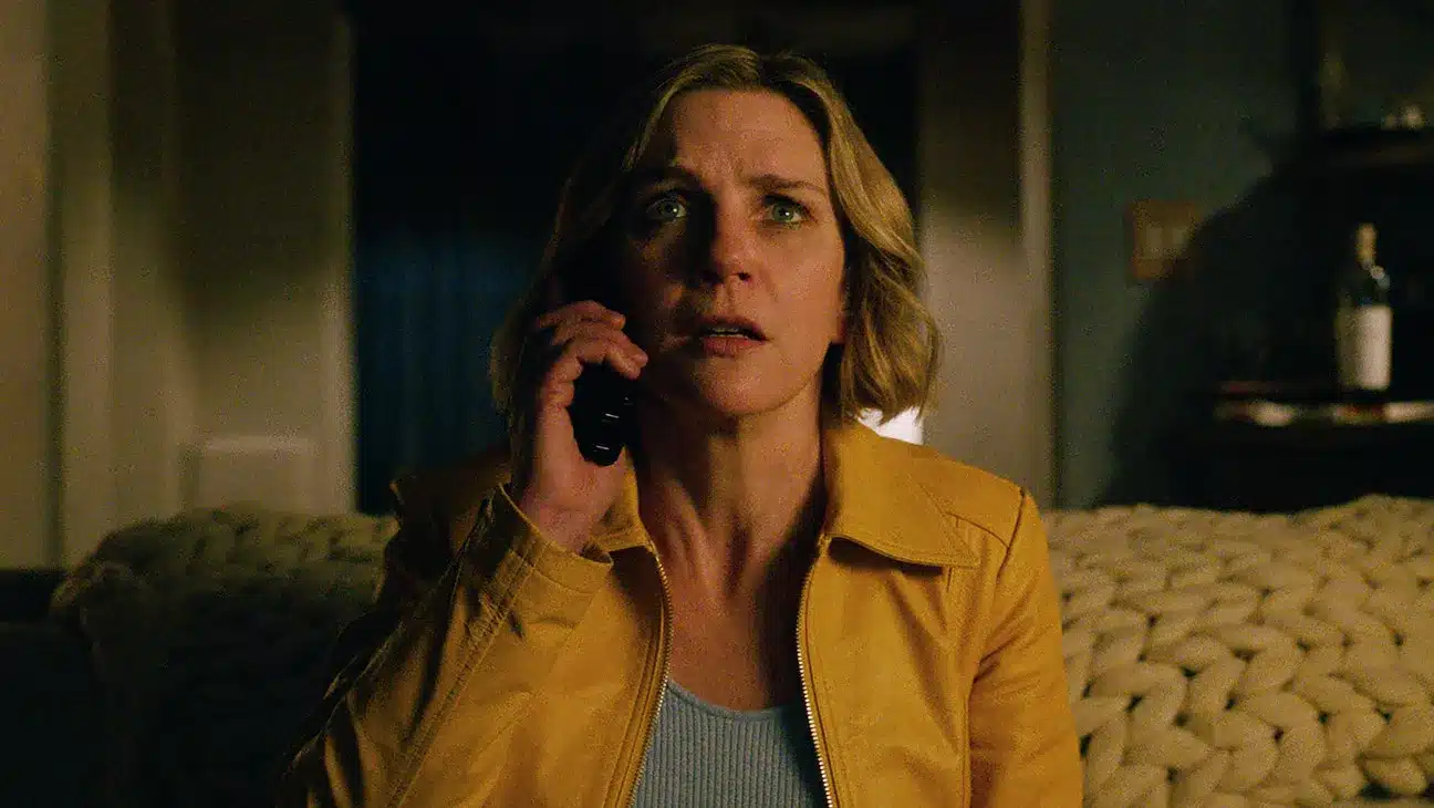 A woman in a yellow jacket looks tense while speaking on the phone in a dimly lit room.