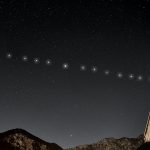 A line of bright Starlink satellites moving across a starry night sky above mountains and a wooden roof.