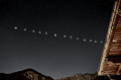 A line of bright Starlink satellites moving across a starry night sky above mountains and a wooden roof.