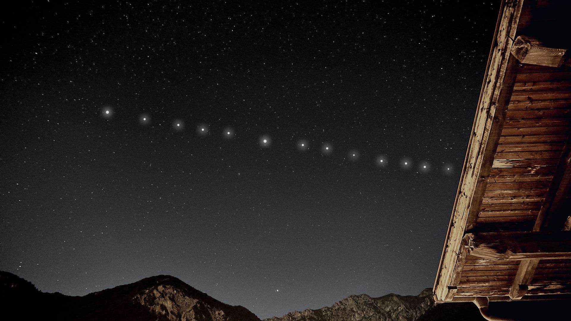 A line of bright Starlink satellites moving across a starry night sky above mountains and a wooden roof.
