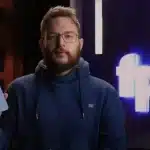 A man in a blue hoodie holds a smartphone while speaking on camera.