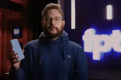A man in a blue hoodie holds a smartphone while speaking on camera.