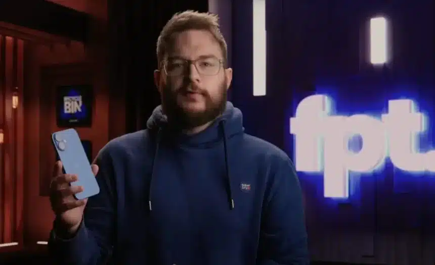 A man in a blue hoodie holds a smartphone while speaking on camera.