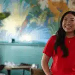 Awkwafina smiling while standing in a colourful restaurant setting.
