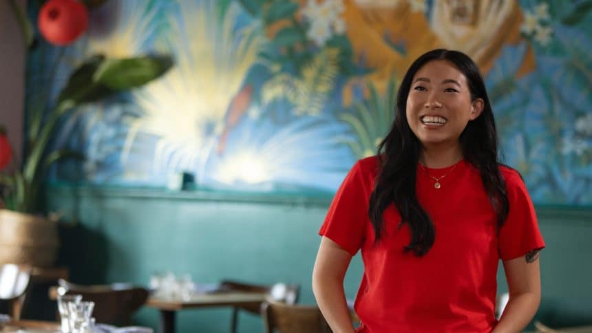 Awkwafina smiling while standing in a colourful restaurant setting.