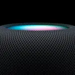 Close-up of Apple’s HomePod mini speaker glowing with colourful light on top.