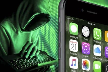 A hacker in a hoodie typing on a laptop next to an iPhone screen.