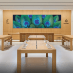 Apple Noida store interior with wooden tables, product displays, and a central video wall.