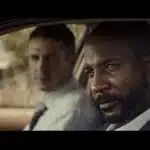 Two men sit inside a car, looking serious during a scene from a video ad.