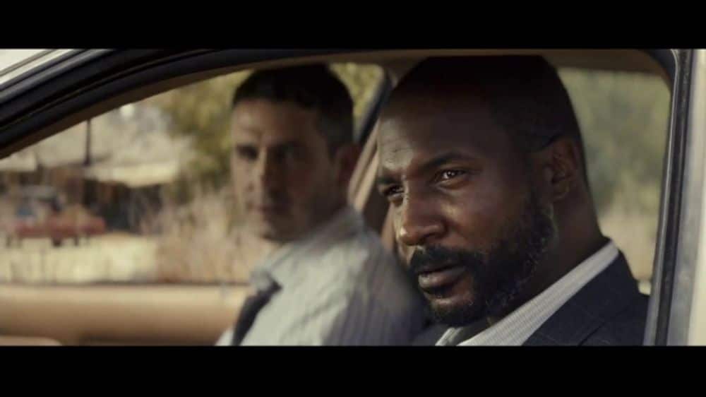 Two men sit inside a car, looking serious during a scene from a video ad.