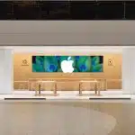 Apple retail store interior with wooden tables, large Apple logo display, and Genius Bar area.