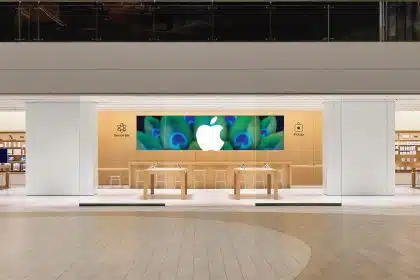 Apple retail store interior with wooden tables, large Apple logo display, and Genius Bar area.