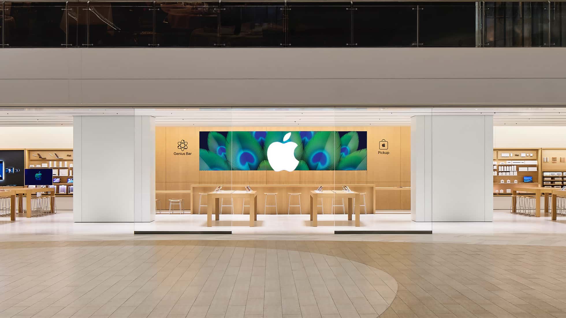 Apple retail store interior with wooden tables, large Apple logo display, and Genius Bar area.