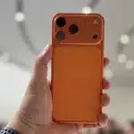 Hand holding an orange iPhone with a multi-camera module in a bright indoor space.