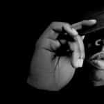 Black-and-white portrait of a man adjusting the brim of his cap against a dark background.